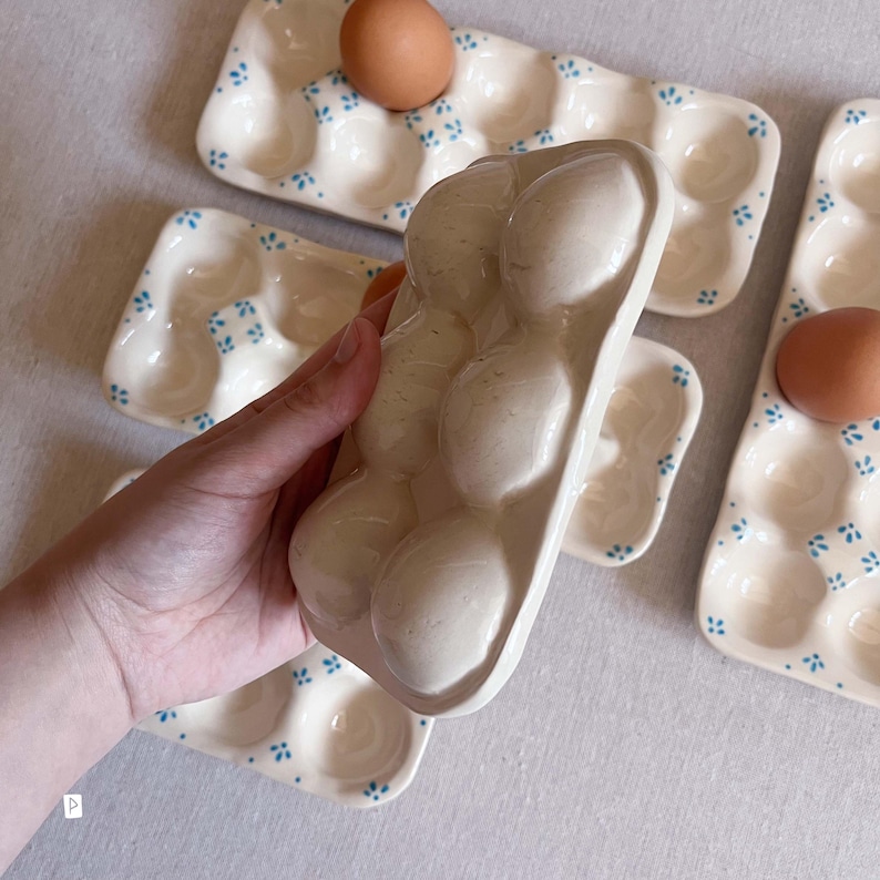 May include: A rectangular ceramic egg tray with a cream-coloured glaze and small blue floral patterns. The tray features six rounded indentations designed to hold eggs securely. Several identical trays are arranged around a central held tray.