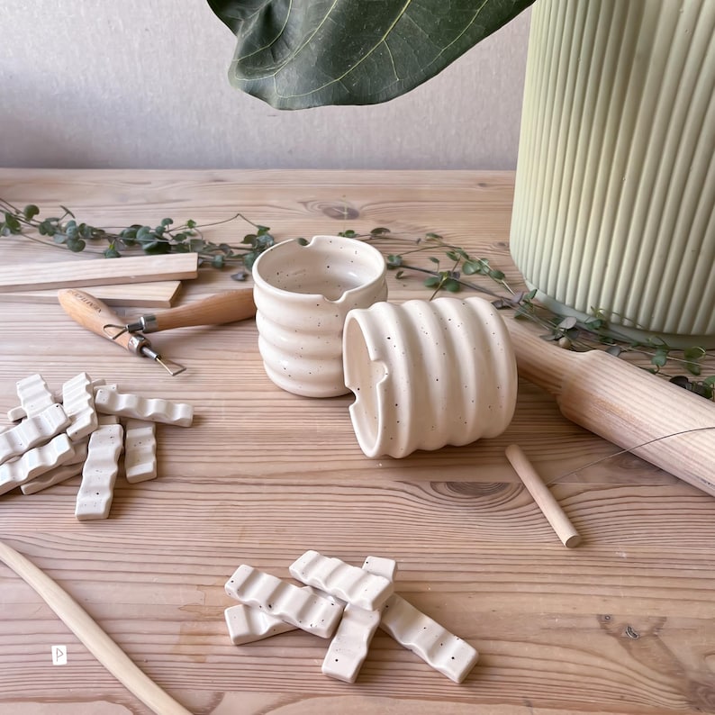 May include: Two white ceramic cups with a speckled glaze and a wavy texture. The cups are sitting on a wooden surface with other ceramic pieces and tools.