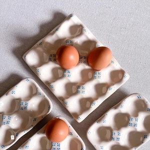 May include: Three white ceramic egg trays with a repeating blue floral pattern. The trays feature moulded indentations for holding eggs. Two brown eggs are placed in the larger central tray, and one brown egg sits in a smaller tray.