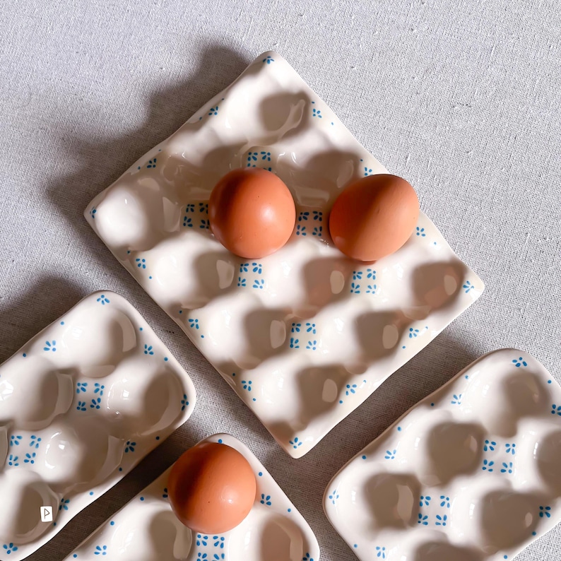May include: Three white ceramic egg trays with a repeating blue floral pattern. The trays feature moulded indentations for holding eggs. Two brown eggs are placed in the larger central tray, and one brown egg sits in a smaller tray.