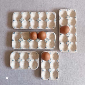 May include: A collection of five ceramic egg trays in varying sizes, featuring a cream-coloured glaze with small blue floral patterns between the indentations. Two trays hold brown eggs, while three trays remain empty, showcasing the glossy finish.