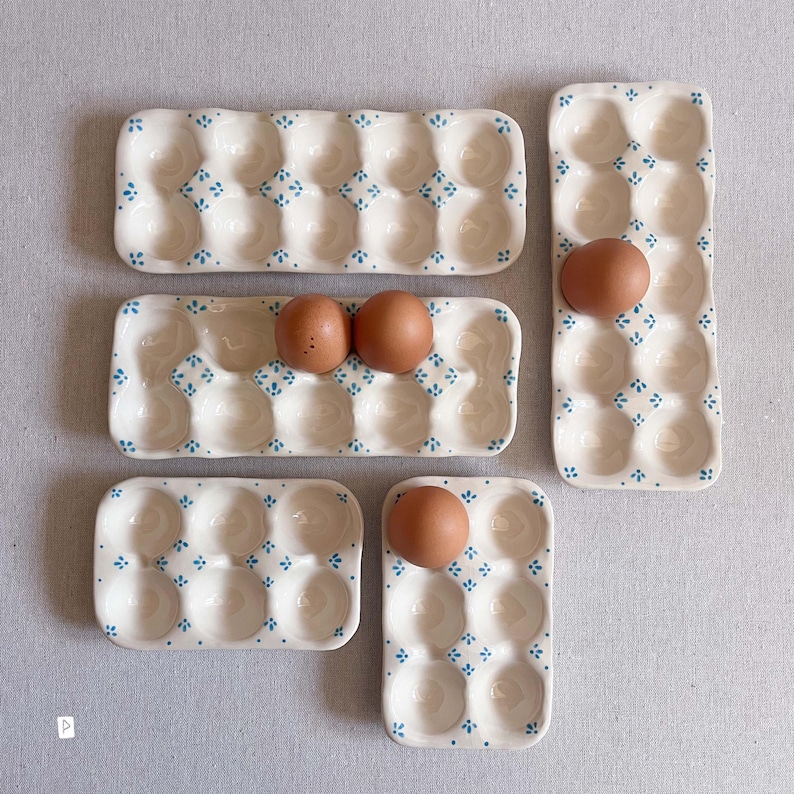 May include: A collection of five ceramic egg trays in varying sizes, featuring a cream-coloured glaze with small blue floral patterns between the indentations. Two trays hold brown eggs, while three trays remain empty, showcasing the glossy finish.