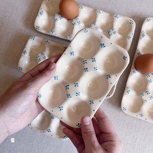 May include: A rectangular ceramic egg tray with a cream-coloured glaze and a repeating pattern of small blue floral motifs. The tray features six circular indentations for holding eggs, with one brown egg resting in a corner slot.