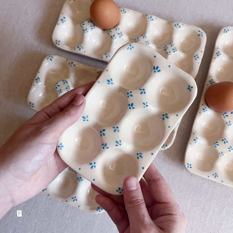 May include: A rectangular ceramic egg tray with a cream-coloured glaze and a repeating pattern of small blue floral motifs. The tray features six circular indentations for holding eggs, with one brown egg resting in a corner slot.