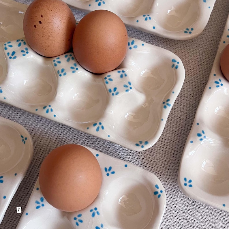 May include: A white ceramic egg tray with six rounded indentations, decorated with small blue painted floral patterns. The tray holds two brown eggs in the top section and one brown egg in the bottom section, with additional empty trays visible.