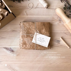 May include: A brown paper package with a white tag that says "Say hi to your new favorite!" and "Recycle the rest of the packaging". The tag is tied to the package with twine. The package is sitting on a wooden surface.