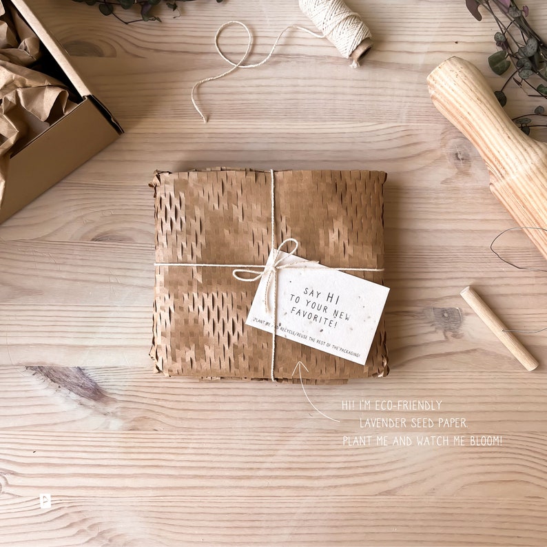 May include: A brown paper package with a white tag that says "Say hi to your new favorite!" and "Recycle the rest of the packaging". The tag is tied to the package with twine. The package is sitting on a wooden surface.