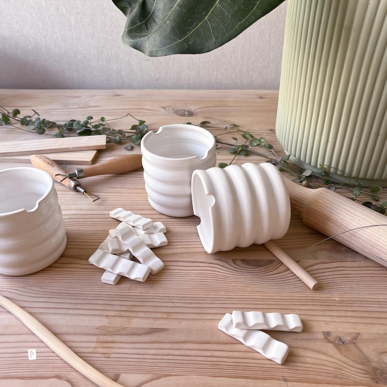 May include: Three white ceramic cups with a wavy, ribbed design. The cups are arranged on a wooden surface with wooden tools and a rolling pin. There are also several white ceramic pieces that look like tiles.