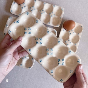May include: A rectangular ceramic egg tray with a cream-coloured glaze and a repeating pattern of small blue floral motifs. The tray features twelve circular indentations for holding eggs, with some brown eggs placed in the slots.