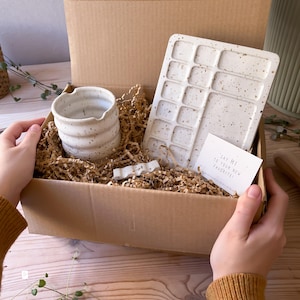 May include: A gift box filled with speckled white ceramic items, including a small mug, a rectangular tray with multiple compartments, and a small ceramic object. The box is filled with brown packing paper and a card that reads "Say Hi to your new favorite!"