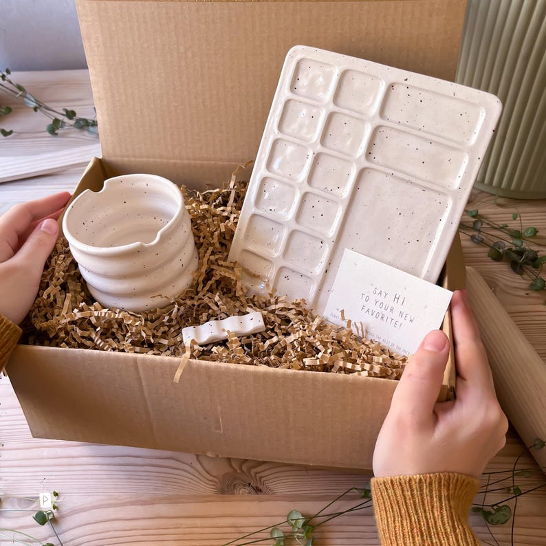 May include: A gift box filled with speckled white ceramic items, including a small bowl, a rectangular palette with multiple wells, and a small, wavy ceramic piece. The box is filled with brown packing paper and a card that reads "Say hi to your new favorite!"