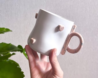 Può includere: Una tazza in ceramica bianca con una superficie liscia e leggermente irregolare, caratterizzata da piccoli cuori in rilievo di colore rosa chiaro. La tazza presenta un manico rotondo rosa chiaro con macchioline blu e nere.