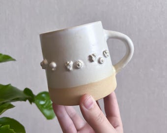 May include: A ceramic mug with a smooth white glazed upper section and a textured, unglazed tan base. The mug features a horizontal row of small, raised, white sculpted shapes encircling the middle, including flowers and abstract forms.