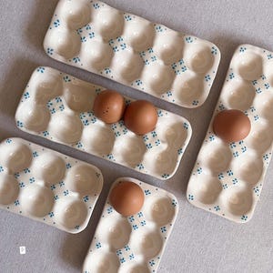 May include: A collection of cream-coloured ceramic egg trays featuring small blue painted floral accents. The trays vary in size, with some holding six eggs and others holding twelve. Several brown eggs are placed within the individual indentations.