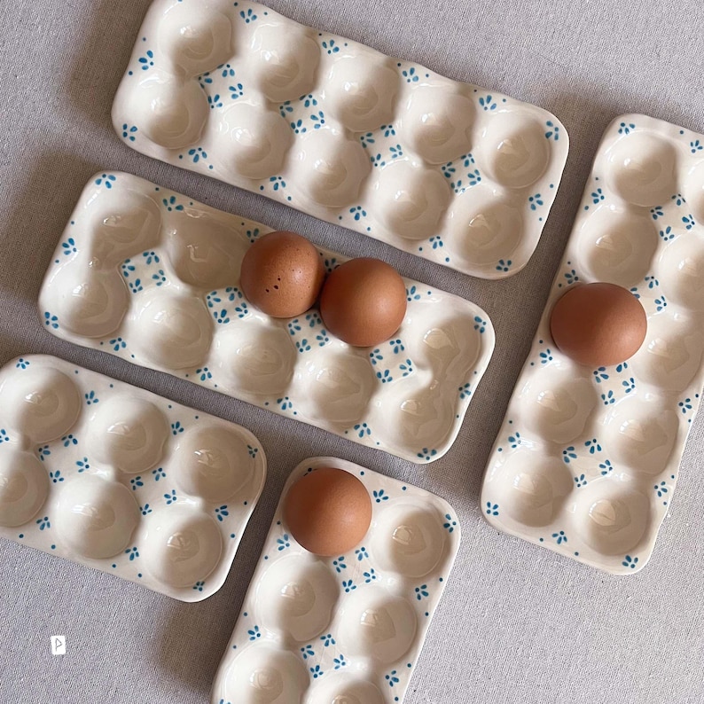 May include: A collection of cream-coloured ceramic egg trays featuring small blue painted floral accents. The trays vary in size, with some holding six eggs and others holding twelve. Several brown eggs are placed within the individual indentations.
