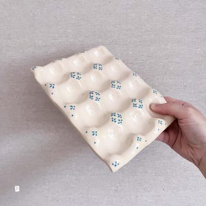 May include: A rectangular ceramic egg tray with a glossy cream-coloured finish, featuring twelve moulded indentations for holding eggs. The surface is decorated with small, hand-painted blue floral patterns scattered between the egg cups.