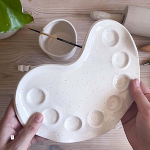 Op de afbeelding: Een wit keramisch verfpalet met een gespikkeld patroon van kleine oranje, gele en zwarte stippen. Het palet heeft zes ronde uitsparingen voor verf en een gladde, gebogen rand. Een kleine keramische waterbeker en een penseel liggen in de buurt.