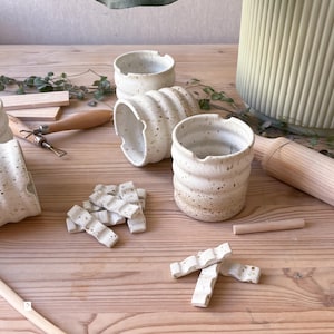 May include: Four white ceramic cups with a speckled glaze and a wavy design. The cups are arranged on a wooden surface with small white ceramic tiles and wooden tools.
