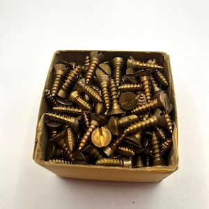May include: A cardboard box filled with numerous brass screws. The screws have slotted heads and are tightly packed together. The box is open, revealing the contents against a white background.