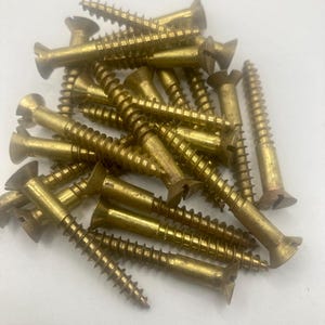 May include: A pile of brass wood screws with slotted heads. The screws have a golden color and are designed for use in woodworking and construction projects. The image shows the screws scattered on a white surface.