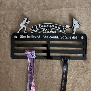 Puede incluir: Un soporte de medallas negro con dos medallas colgadas. El soporte tiene una silueta blanca de una jugadora de tenis a cada lado. El texto "Champion Alisha" est&aacute; en la parte superior del soporte. El texto "She believed, She could, So She did" est&aacute; debajo del nombre. La medalla de la izquierda es morada y blanca con el texto "2022" en ella. La medalla de la derecha es negra con texto dorado que dice "TORONTO WATERFRONT MARATHON".