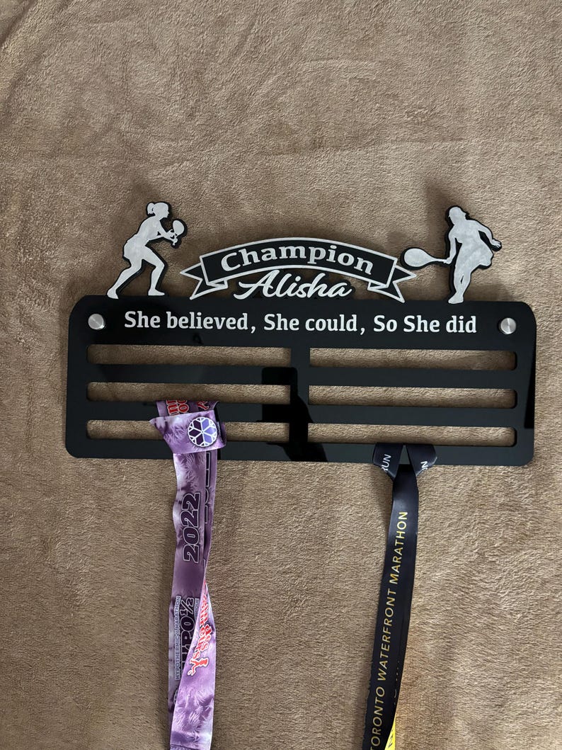 Puede incluir: Un soporte de medallas negro con dos medallas colgadas. El soporte tiene una silueta blanca de una jugadora de tenis a cada lado. El texto "Champion Alisha" est&aacute; en la parte superior del soporte. El texto "She believed, She could, So She did" est&aacute; debajo del nombre. La medalla de la izquierda es morada y blanca con el texto "2022" en ella. La medalla de la derecha es negra con texto dorado que dice "TORONTO WATERFRONT MARATHON".