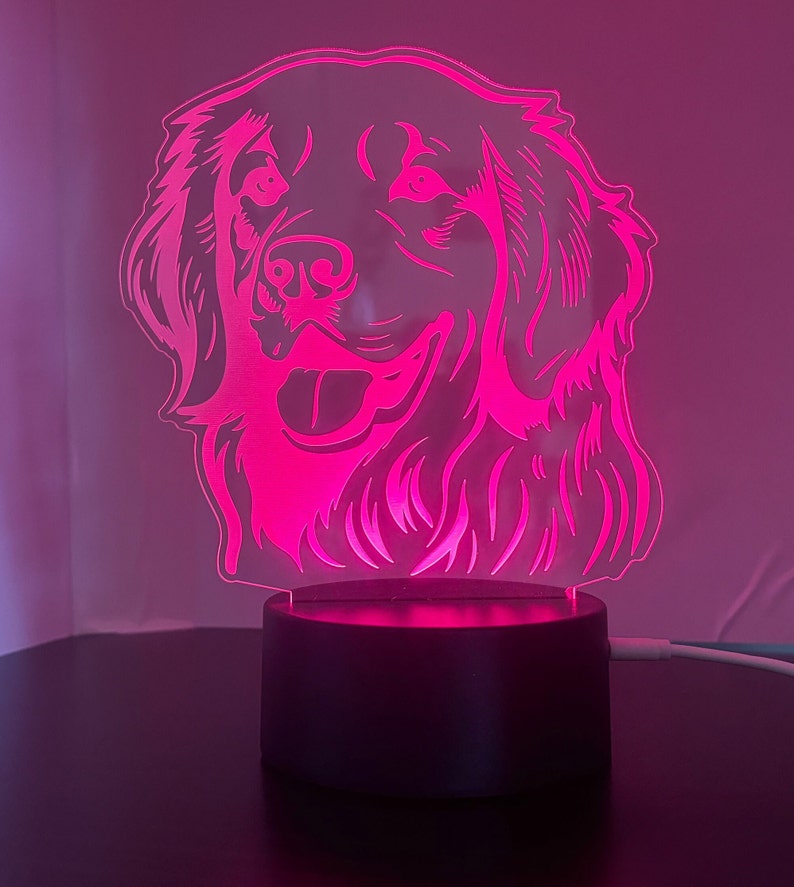 May include: A pink acrylic night light shaped like a golden retriever dog's head. The light is on a black base.