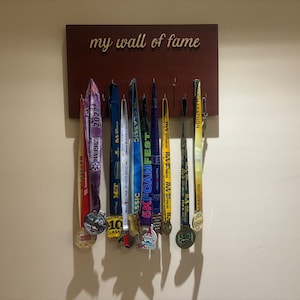 Puede incluir: Una placa de madera con el texto "my wall of fame"  mostrada en una pared con una colección de medallas coloridas colgadas de ganchos.