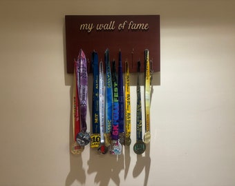 Puede incluir: Una placa de madera con el texto "my wall of fame"  mostrada en una pared con una colección de medallas coloridas colgadas de ganchos.