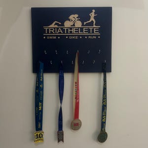 Puede incluir: Un tablero de madera azul para exhibir medallas con el texto "TRIATHELETE" y "* SWIM * * BIKE * * RUN *" en letras doradas. El tablero tiene ganchos para colgar medallas. Hay tres medallas colgadas en el tablero, una roja, una azul y una amarilla.