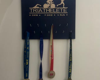 May include: A blue wooden medal display board with the text "TRIATHELETE" and "* SWIM * * BIKE * * RUN *" in gold lettering. The board has hooks for hanging medals. Three medals are hanging on the board, one red, one blue, and one yellow.