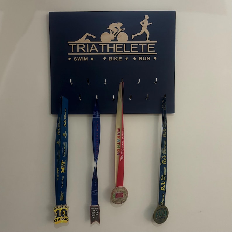 K&ouml;nnte beinhalten: Ein blaues, h&ouml;lzernes Medaillen-Pr&auml;sentationsbrett mit dem Text "TRIATHELETE" und "* SWIM * * BIKE * * RUN *" in goldenen Buchstaben. Das Brett hat Haken zum Aufh&auml;ngen von Medaillen. Drei Medaillen h&auml;ngen am Brett, eine rote, eine blaue und eine gelbe.