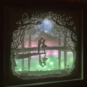 K&ouml;nnte beinhalten: Ein wei&szlig;es Papier-Schnittbild, das ein Paar zeigt, das an einem See in einem Wald steht. Das Paar ist vor einem mondbeschienenen Himmel mit Sternen silhouettiert. Der See spiegelt den Mond und das Paar wider.
