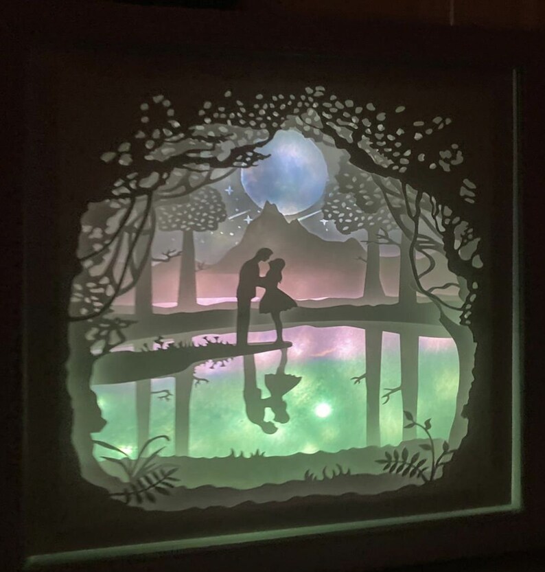 K&ouml;nnte beinhalten: Ein wei&szlig;es Papier-Schnittbild, das ein Paar zeigt, das an einem See in einem Wald steht. Das Paar ist vor einem mondbeschienenen Himmel mit Sternen silhouettiert. Der See spiegelt den Mond und das Paar wider.