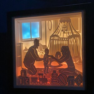 Op de afbeelding: Een schaduwdoos van papier met een gezinsscene. Een stel zit op de vloer met hun baby die tussen hen staat. De baby reikt naar de moeder. De sc&egrave;ne is van achteren verlicht, waardoor een warme gloed ontstaat. De doos bevat een wieg, speelgoed en een raam met een blauwe lucht.