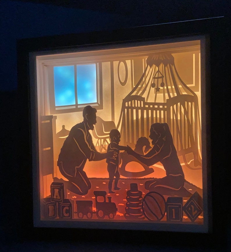 Op de afbeelding: Een schaduwdoos van papier met een gezinsscene. Een stel zit op de vloer met hun baby die tussen hen staat. De baby reikt naar de moeder. De sc&egrave;ne is van achteren verlicht, waardoor een warme gloed ontstaat. De doos bevat een wieg, speelgoed en een raam met een blauwe lucht.