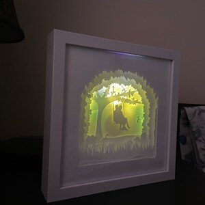 K&ouml;nnte beinhalten: Eine wei&szlig;e Schattenbox mit einem wei&szlig;en Rahmen, die eine Papierschnitt-Gestaltung eines Paares zeigt, das auf einer Schaukel unter einem Baum sitzt. Das Design wird von hinten mit einem warmen gelben Licht beleuchtet.