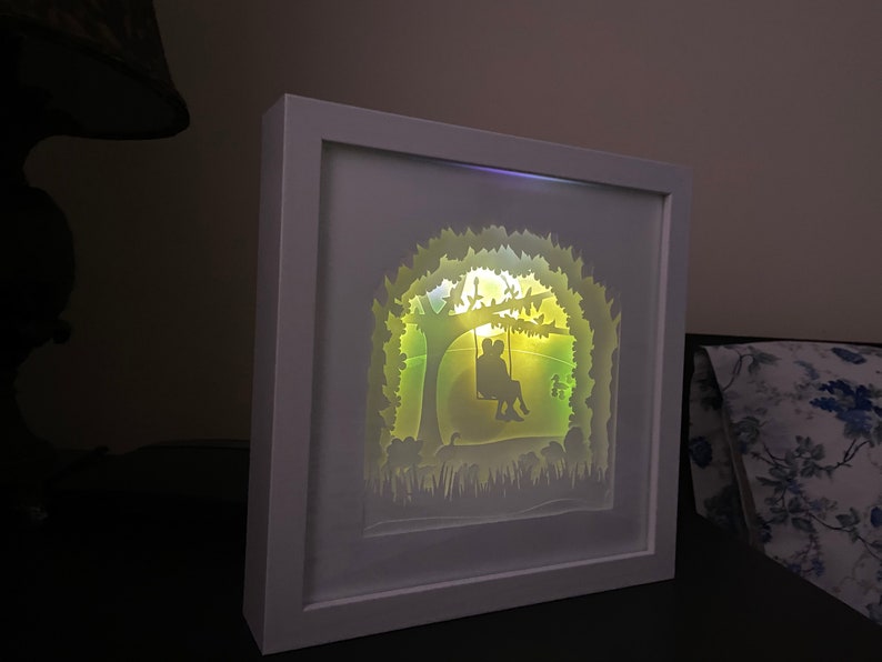 K&ouml;nnte beinhalten: Eine wei&szlig;e Schattenbox mit einem wei&szlig;en Rahmen, die eine Papierschnitt-Gestaltung eines Paares zeigt, das auf einer Schaukel unter einem Baum sitzt. Das Design wird von hinten mit einem warmen gelben Licht beleuchtet.