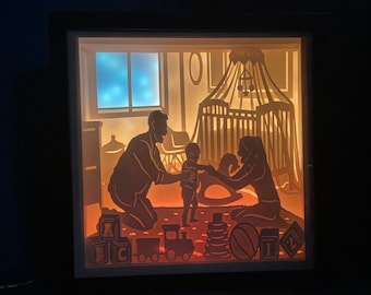 Puede incluir: Una caja de luz con una silueta de una familia en una habitación de bebé. El padre sostiene un bebé y la madre está sentada en el suelo. La habitación de bebé tiene una cuna, un caballo de balancín y juguetes.