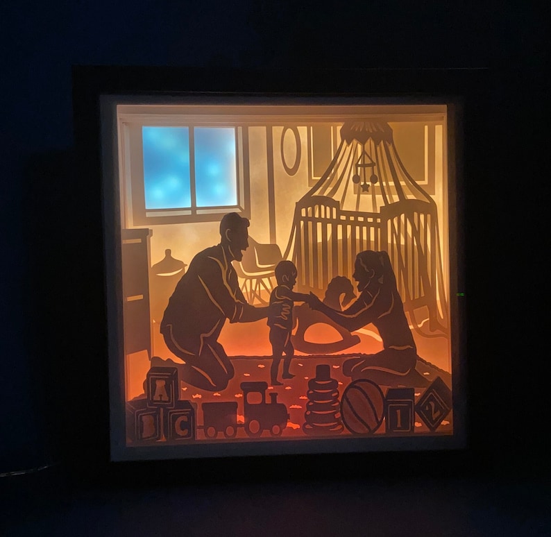 Op de afbeelding: Een schaduwdoos met een silhouet van een gezin in een kinderkamer. De vader houdt een baby vast en de moeder zit op de grond. De kinderkamer heeft een wieg, een schommelpaard en speelgoed.