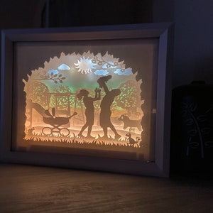 K&ouml;nnte beinhalten: Eine wei&szlig;e Schattenbox mit einem Papierschnitt-Design einer Familie in einem Park. Die Familie ist vor einem gr&uuml;n-blauen Hintergrund mit Sonne und Wolken silhouettiert. Das Design beinhaltet einen Kinderwagen, B&auml;ume und ein kleines Tier.