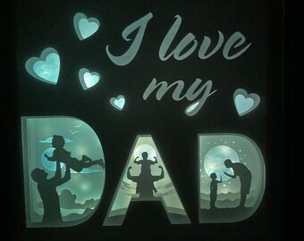 Puede incluir: Un letrero blanco iluminado con las palabras "I love my DAD" en una fuente decorativa. El letrero presenta tres escenas en silueta de un padre y un hijo, con una luna y estrellas en el fondo.
