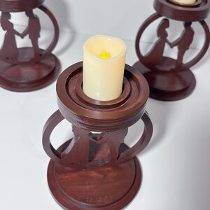 K&ouml;nnte beinhalten: Drei dunkelbraune Kerzenhalter aus Holz. Jeder Halter zeigt eine Silhouette eines Paares, das sich an den H&auml;nden h&auml;lt. Die Halter haben einen kreisf&ouml;rmigen Sockel und ein Ringdesign. Cremefarbene LED-Kerzen sind in jedem Halter.