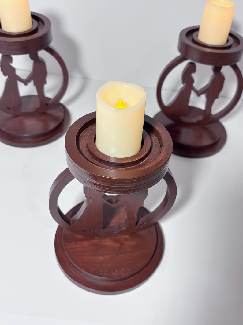 K&ouml;nnte beinhalten: Drei dunkelbraune Kerzenhalter aus Holz. Jeder Halter zeigt eine Silhouette eines Paares, das sich an den H&auml;nden h&auml;lt. Die Halter haben einen kreisf&ouml;rmigen Sockel und ein Ringdesign. Cremefarbene LED-Kerzen sind in jedem Halter.