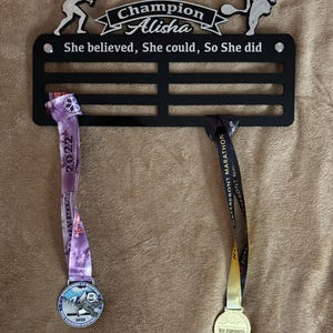Puede incluir: Expositor de medallas de metal negro con dos medallas colgadas. El expositor tiene el texto "Champion Alisha" y "She believed, She could, So She did". Las medallas son plateadas y doradas con cintas.