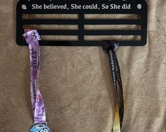 Puede incluir: Expositor de medallas de metal negro con dos medallas colgadas. El expositor tiene el texto "Champion Alisha" y "She believed, She could, So She did". Las medallas son plateadas y doradas con cintas.