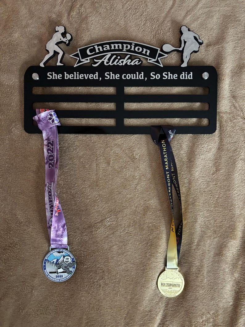 Puede incluir: Expositor de medallas de metal negro con dos medallas colgadas. El expositor tiene el texto "Champion Alisha" y "She believed, She could, So She did". Las medallas son plateadas y doradas con cintas.