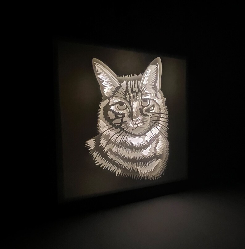 May include: A black and white illustration of a cat's face, illuminated from behind, creating a shadow box effect.