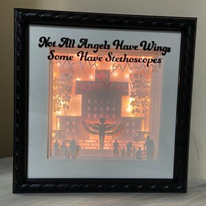 May include: A black framed shadow box with a white background and a paper cut-out design of a hospital building with the text "Not All Angels Have Wings Some Have Stethoscopes". The hospital building has a red sign that says "Hospital" and a lit-up window with the word "Emergency". There are paper cut-out figures of people standing in front of the hospital.