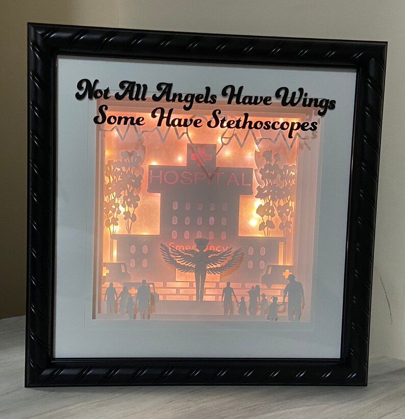 May include: A black framed shadow box with a white background and a paper cut-out design of a hospital building with the text "Not All Angels Have Wings Some Have Stethoscopes". The hospital building has a red sign that says "Hospital" and a lit-up window with the word "Emergency". There are paper cut-out figures of people standing in front of the hospital.